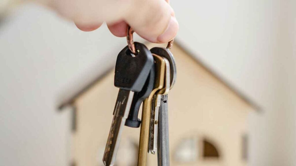 Hand holding house keys in front of a wooden house model representing housing costs in a bare minimum budget.